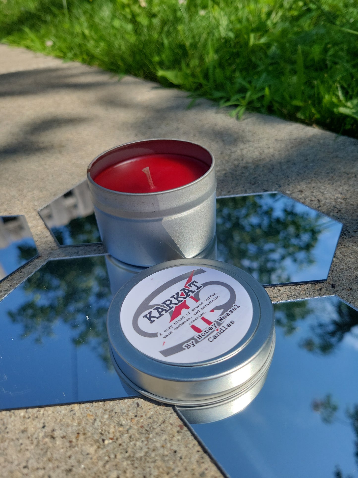A Karkat Vantas Inspired Candle. A silver 4 oz tin with a bright red beeswax candle inside sits on hexagonal shaped mirrors. The lid sits next to it, baring the cancer symbol, the blood aspect symbol, and the title of the candle with a brief description of its scent.