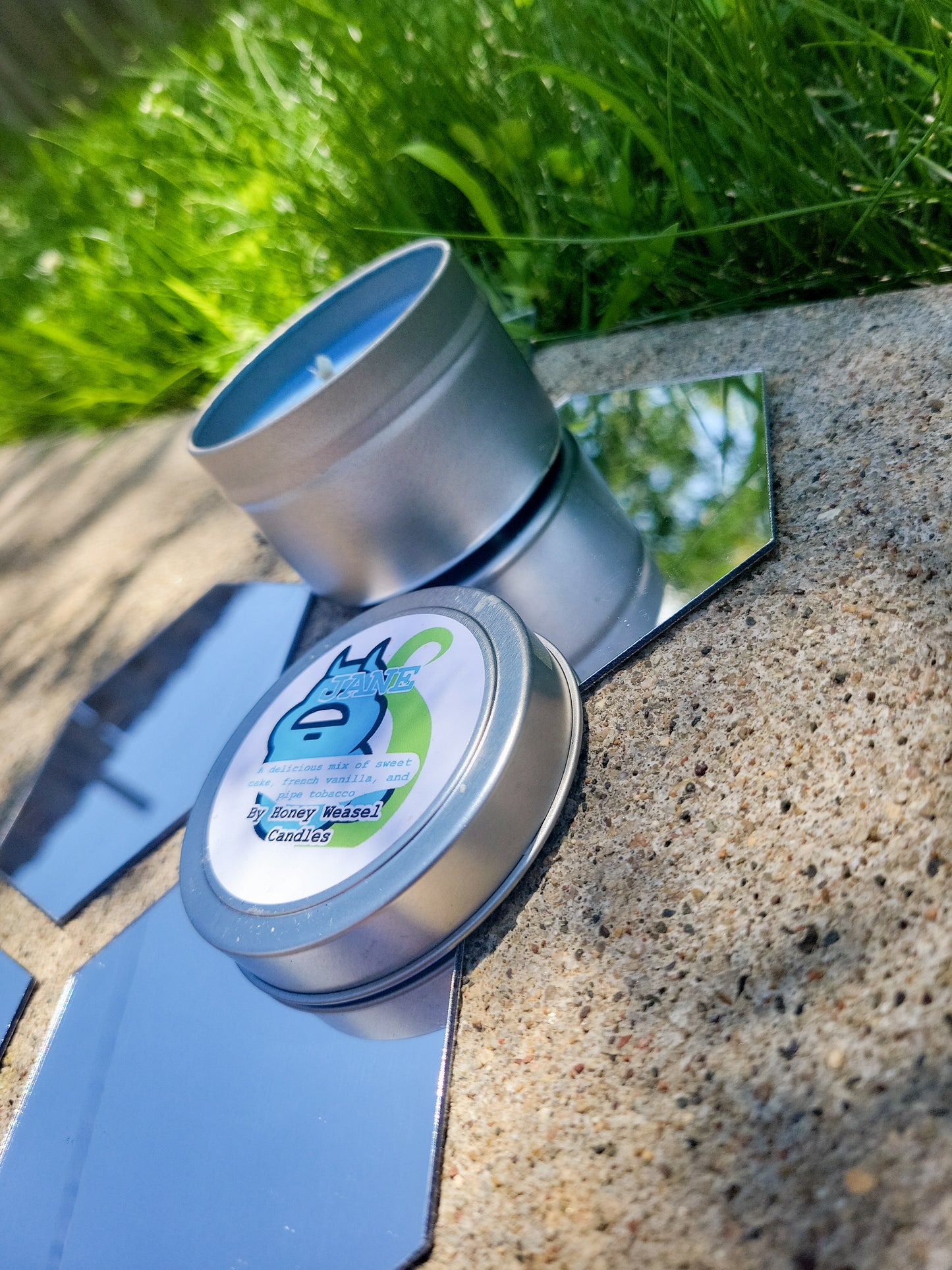 A Jane Crocker Inspired Candle. A silver 4 oz tin with a baby blue beeswax candle inside sits on hexagonal shaped mirrors. The lid sits next to it, baring the blue ghost symbol, the life aspect symbol, and the title of the candle with a brief description of its scent.