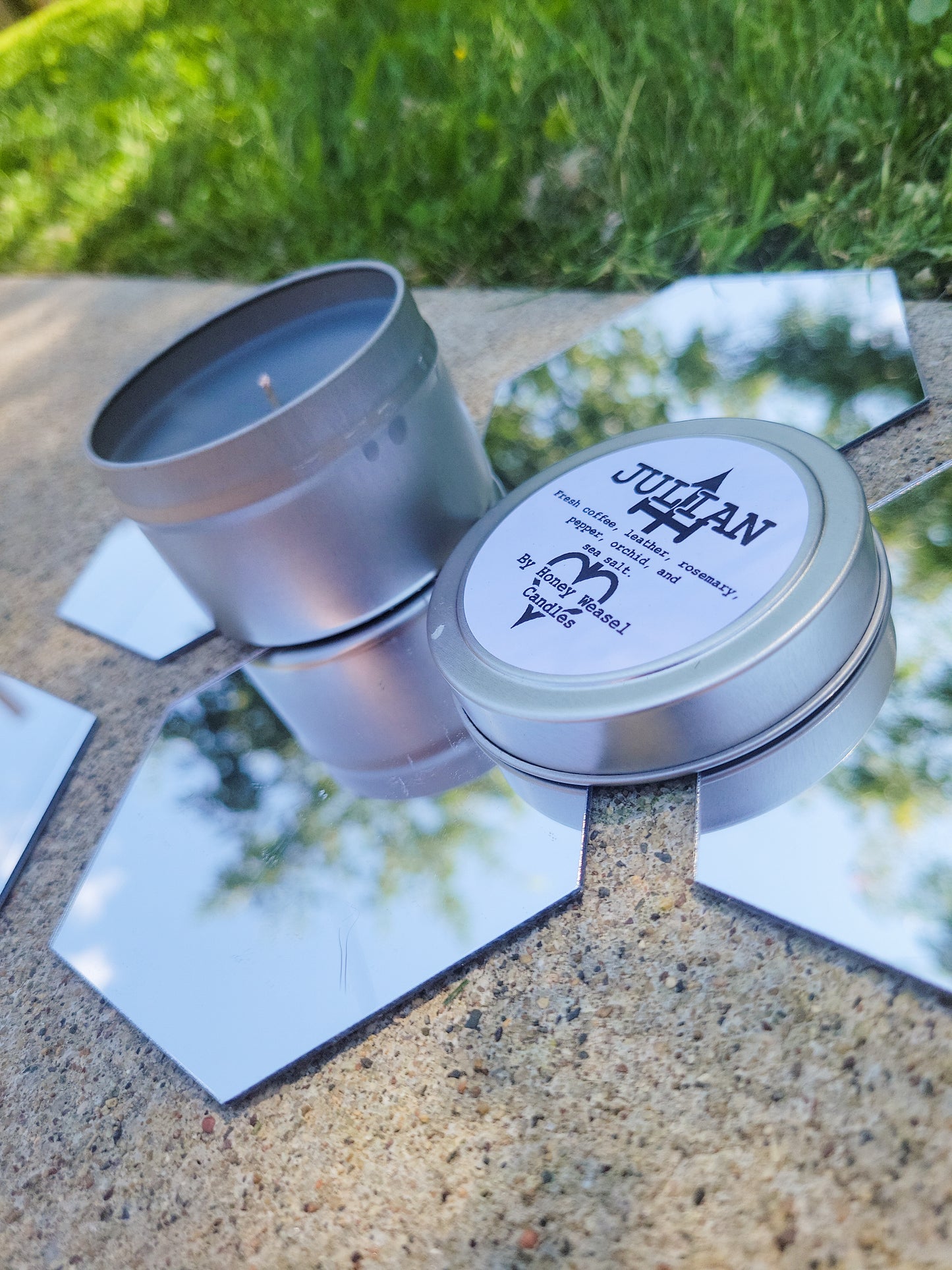 A Julian Devorak Inspired Candle. A silver 4 oz tin with a dark grey beeswax candle inside sits on hexagonal shaped mirrors. The lid sits next to it, baring the "murderer's brand" from The Arcana, and the title of the candle with a brief description of its scent.