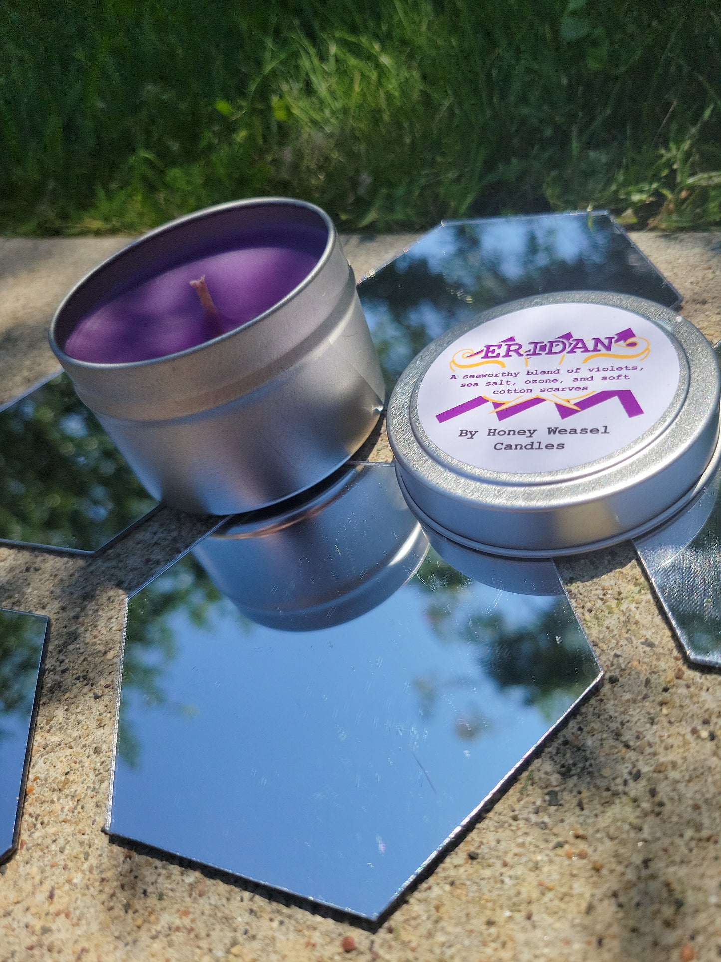 An Eridan Ampora Inspired Candle. A silver 4 oz tin with a bright violet beeswax candle inside sits on hexagonal shaped mirrors. The lid sits next to it, baring the aquarius symbol, the hope aspect symbol, and the title of the candle with a brief description of its scent.