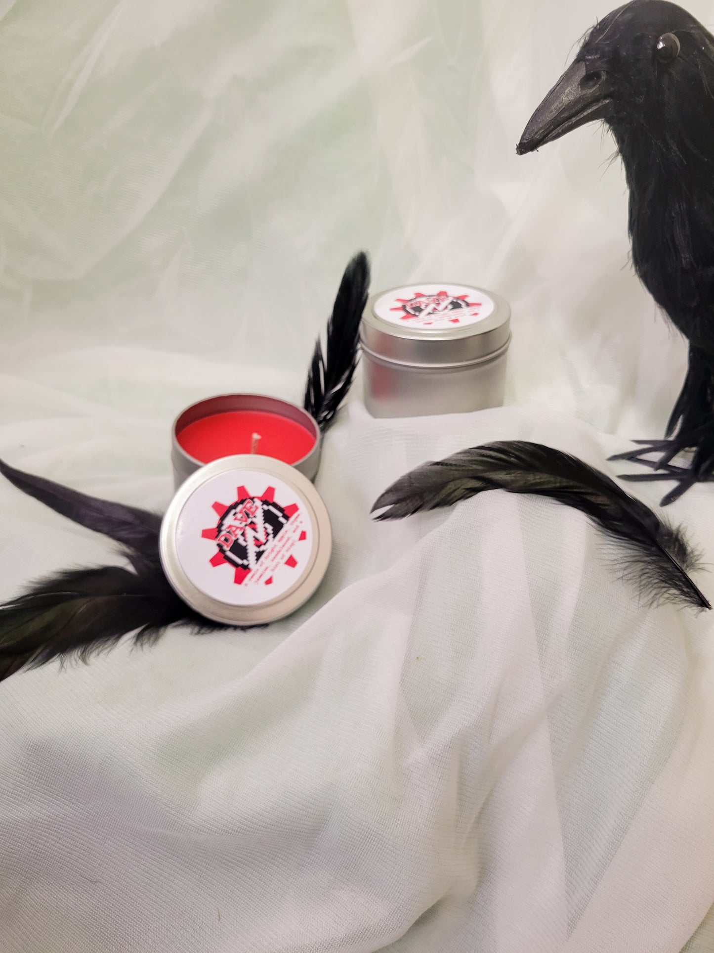 A Dave Strider Inspired Candle. A silver 4 oz tin with a bright red beeswax candle inside sits in white fabric admist black feathers and a fake crow. The lid sits next to it, baring the broken record symbol, the time aspect symbol, and the title of the candle with a brief description of its scent.