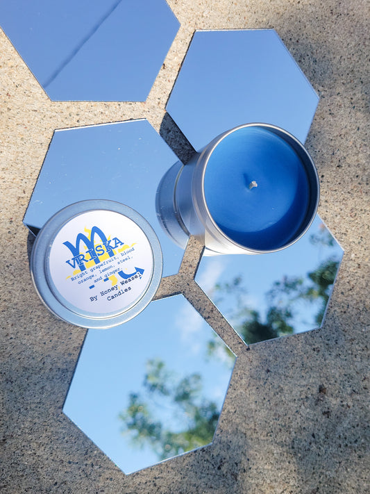 A Vriska Serket Inspired Candle. A silver 4 oz tin with a bright cerulean beeswax candle inside sits on hexagonal shaped mirrors. The lid sits next to it, baring the scorpio symbol, the light aspect symbol, and the title of the candle with a brief description of its scent.