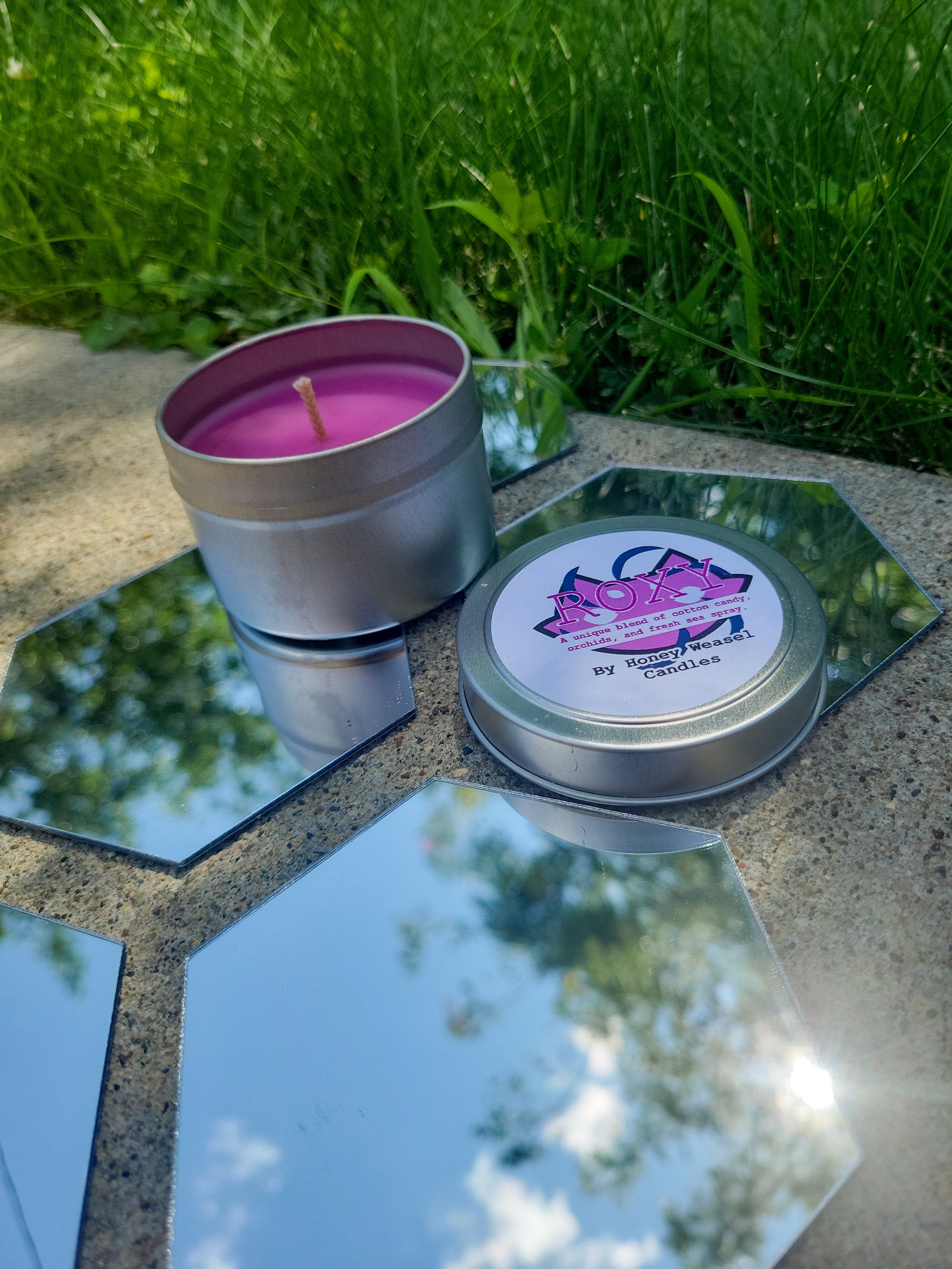 A Roxy Lalonde Inspired Candle. A silver 4 oz tin with a bright pink beeswax candle inside sits on hexagonal shaped mirrors. The lid sits next to it, baring the cat symbol, the void aspect symbol, and the title of the candle with a brief description of its scent.