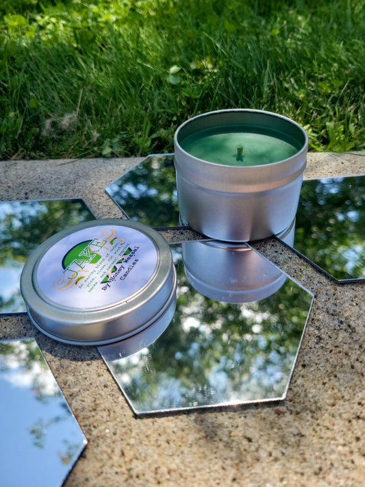 A Jake English Inspired Candle. A silver 4 oz tin with a vivid green beeswax candle inside sits on hexagonal shaped mirrors. The lid sits next to it, baring the green skull symbol, the hope aspect symbol, and the title of the candle with a brief description of its scent.