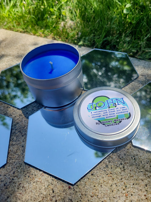 An Egbert Inspired Candle. A silver 4 oz tin with a bright blue beeswax candle inside sits on hexagonal shaped mirrors. The lid sits next to it, baring their slimer symbol, the breath aspect symbol, and the title of the candle with a brief description of its scent.