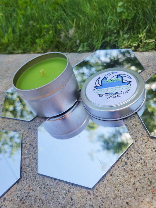 A Jade Harley Inspired Candle. A silver 4 oz tin with a bright green beeswax candle inside sits on hexagonal shaped mirrors. The lid sits next to it, baring her dog symbol, the space aspect symbol, and the title of the candle with a brief description of its scent.