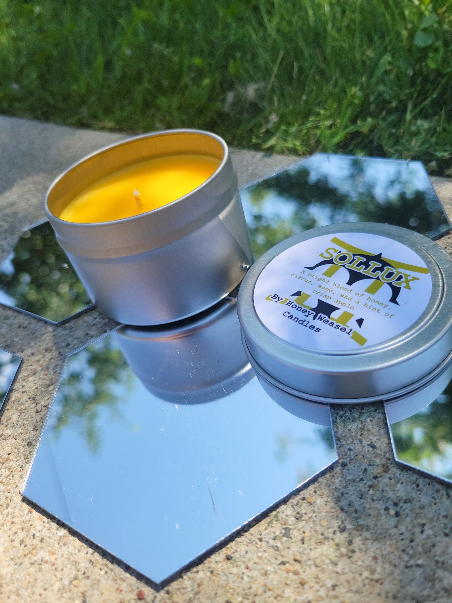 A Sollux Captor Inspired Candle. A silver 4 oz tin with a bright yellow beeswax candle inside sits on hexagonal shaped mirrors. The lid sits next to it, baring the Gemini symbol, the doom aspect symbol, and the title of the candle with a brief description of its scent.