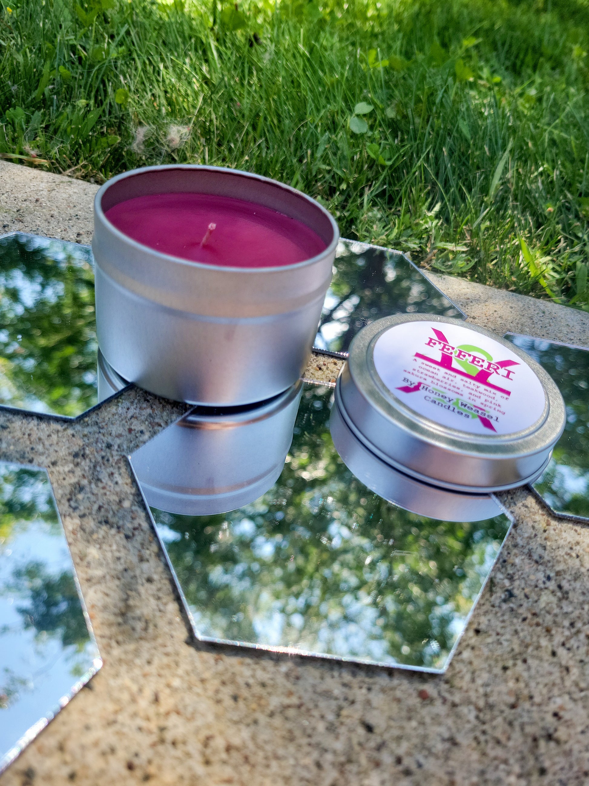 A Feferi Peixes Inspired Candle. A silver 4 oz tin with a bright fushia beeswax candle inside sits on hexagonal shaped mirrors. The lid sits next to it, baring the pisces symbol, the life aspect symbol, and the title of the candle with a brief description of its scent.