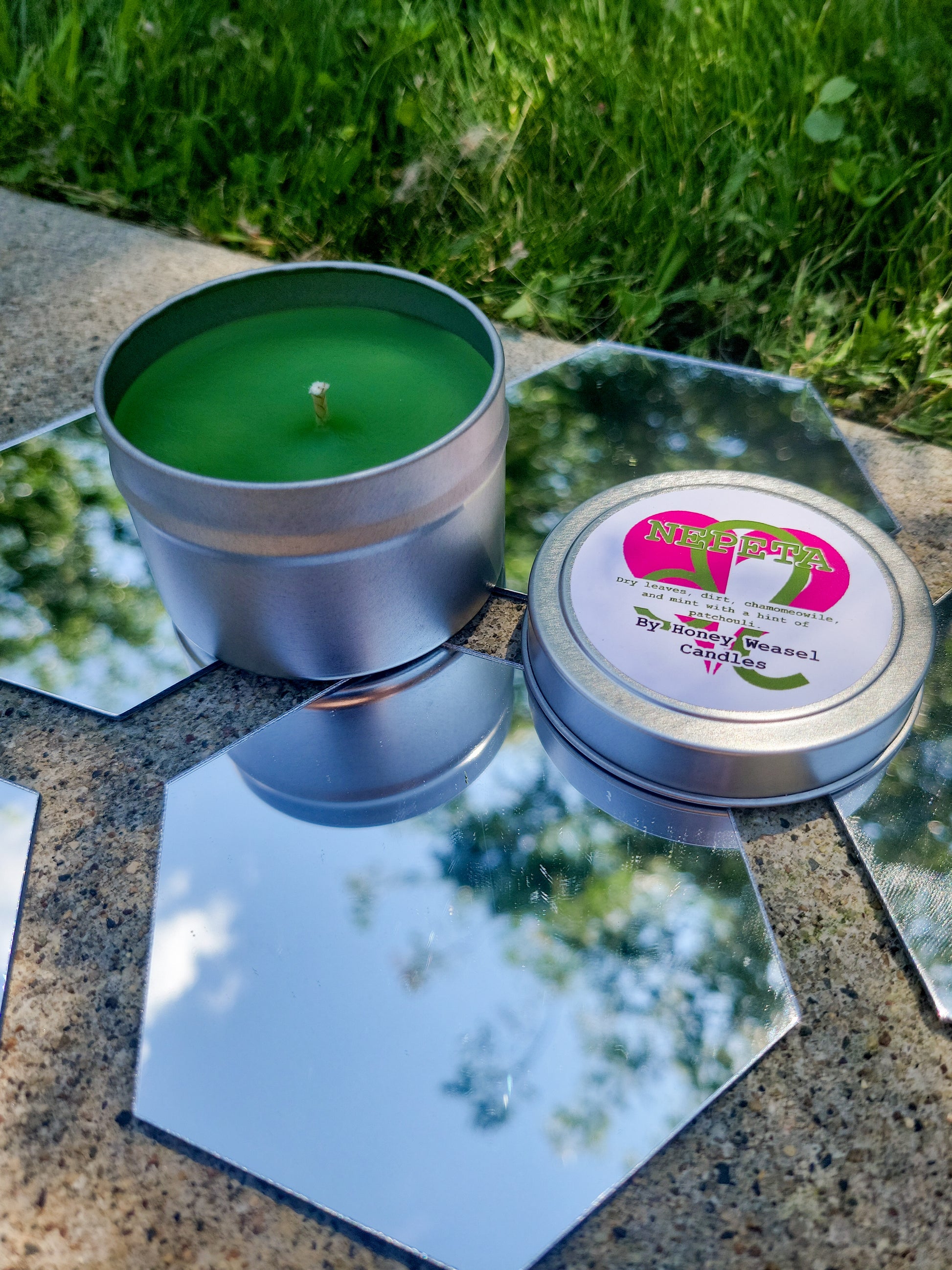 A Nepeta Leijon Inspired Candle. A silver 4 oz tin with an olive green beeswax candle inside sits on hexagonal shaped mirrors. The lid sits next to it, baring the leo symbol, the heart aspect symbol, and the title of the candle with a brief description of its scent.