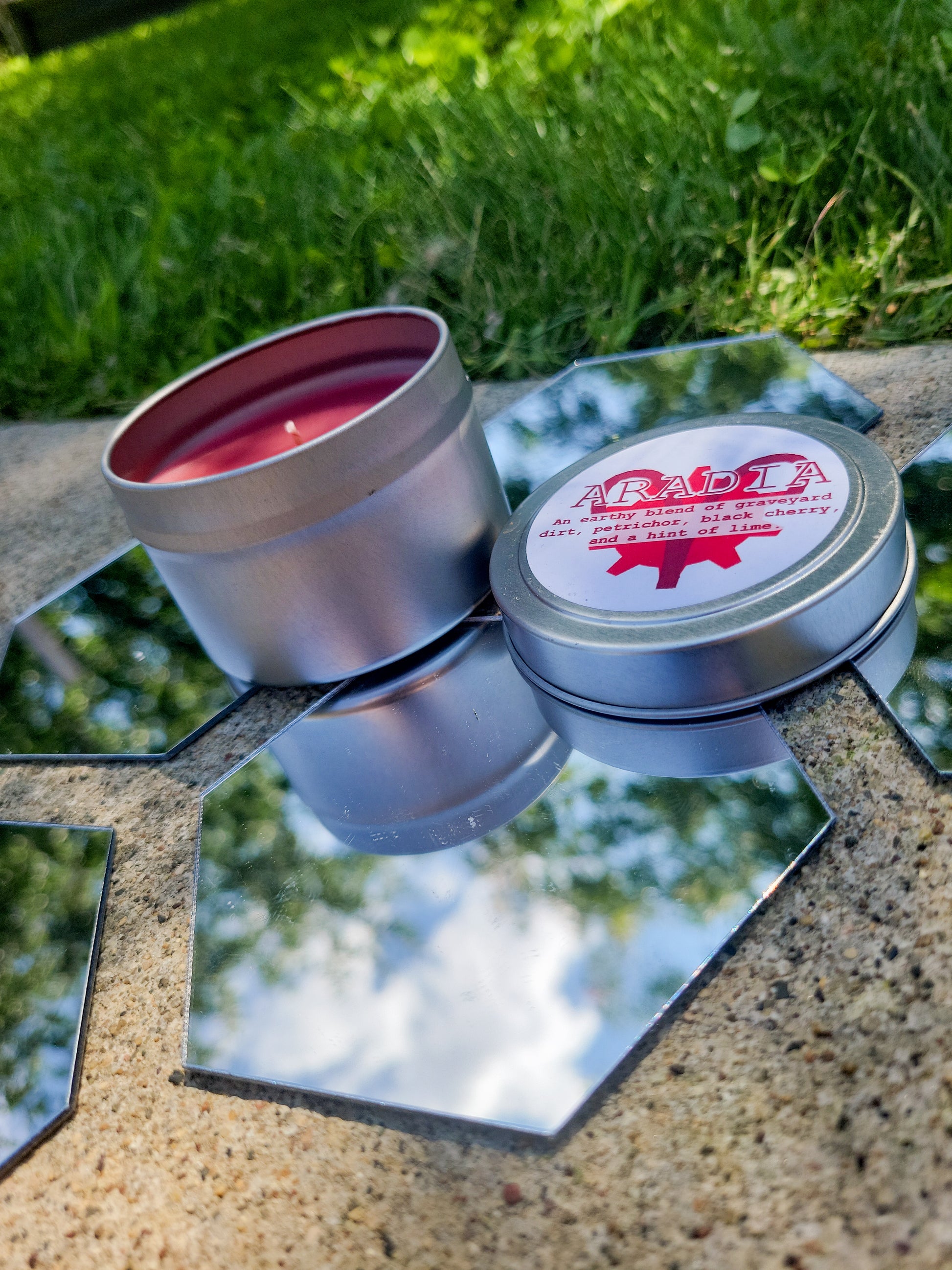 An Aradia Megido Inspired Candle. A silver 4 oz tin with a deep red beeswax candle inside sits on hexagonal shaped mirrors. The lid sits next to it, baring the aries symbol, the time aspect symbol, and the title of the candle with a brief description of its scent.