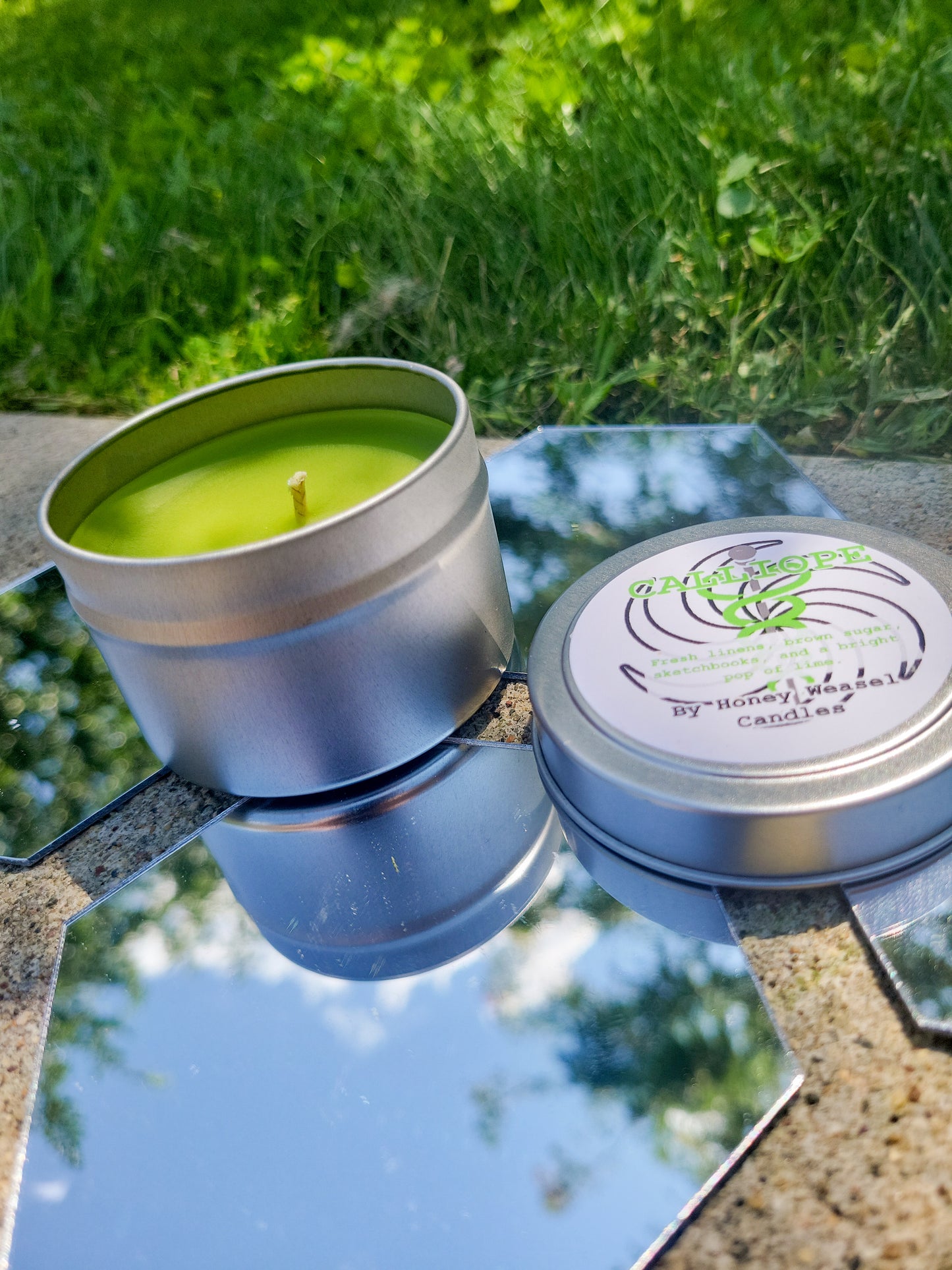 A Calliope Inspired Candle. A silver 4 oz tin with a bright lime green beeswax candle inside sits on hexagonal shaped mirrors. The lid sits next to it, baring the Ophiuchus symbol, the space aspect symbol, and the title of the candle with a brief description of its scent.