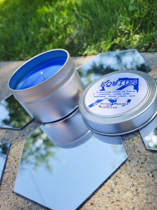 An Equius Zahhak Inspired Candle. A silver 4 oz tin with a bright blue beeswax candle inside sits on hexagonal shaped mirrors. The lid sits next to it, baring the Sagittarius symbol, the void aspect symbol, and the title of the candle with a brief description of its scent.