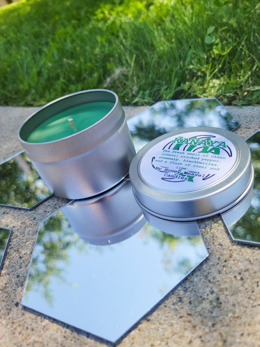 A Kanaya Maryam Inspired Candle. A silver 4 oz tin with a jade green beeswax candle inside sits on hexagonal shaped mirrors. The lid sits next to it, baring the virgo symbol, the space aspect symbol, and the title of the candle with a brief description of its scent.