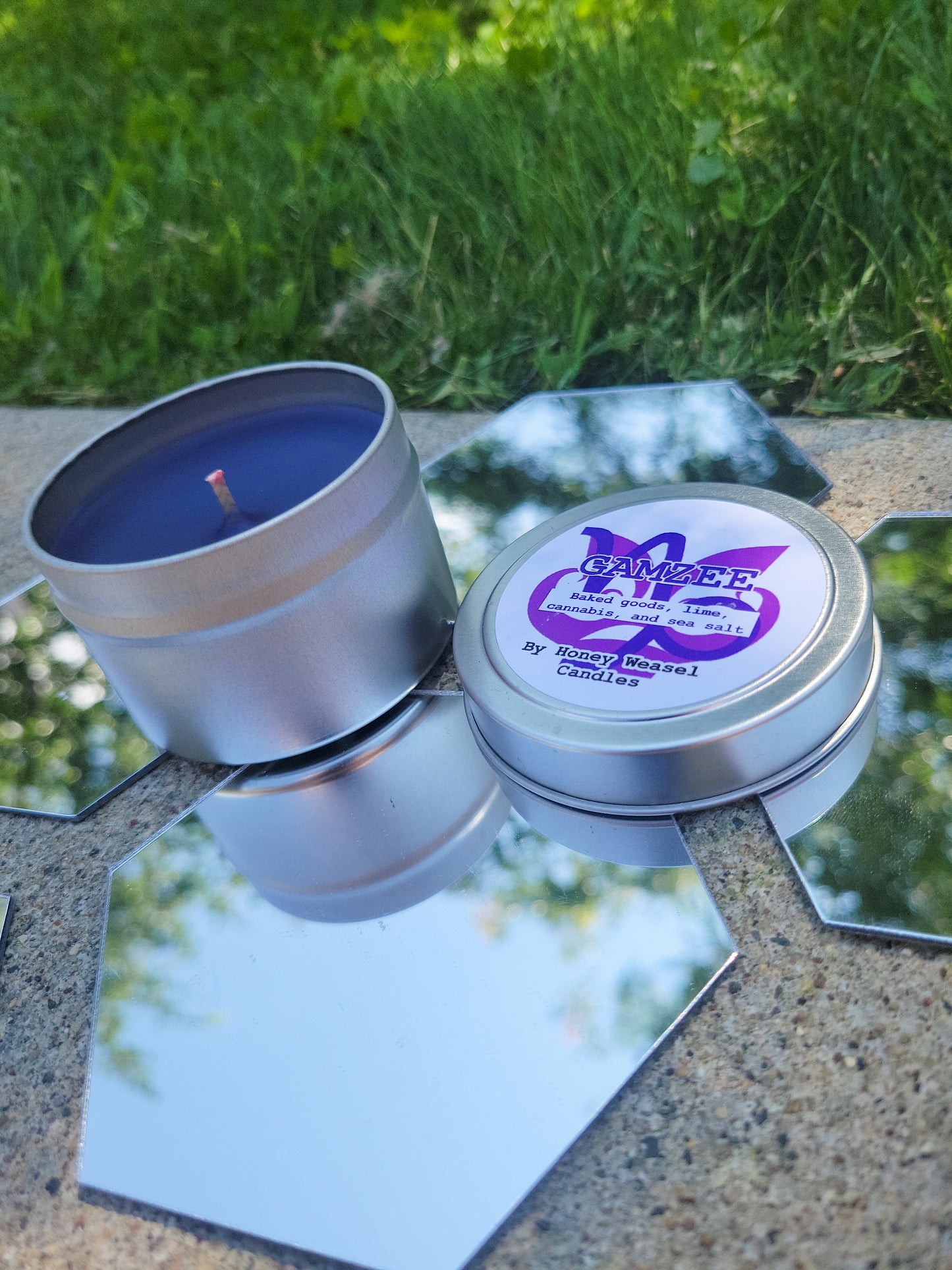 A Gamzee Makara Inspired Candle. A silver 4 oz tin with a deep purple beeswax candle inside sits on hexagonal shaped mirrors. The lid sits next to it, baring the Capricorn symbol, the rage aspect symbol, and the title of the candle with a brief description of its scent.